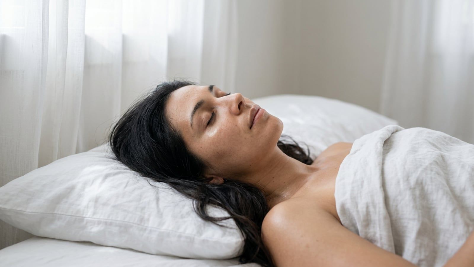 A woman in her thirties asleep on her back on a white cotton pillowcase, early morning light from a window across the bed; olive skin, faint under-eye shadows, hair spread loose.