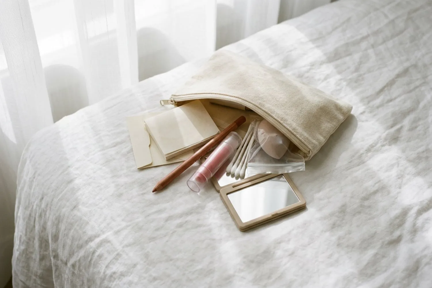 A small zip pouch open on white linen, seen from above: blotting papers, a lip liner, a tube of tinted balm, cotton swabs, a sponge in a clear bag and a folding travel mirror