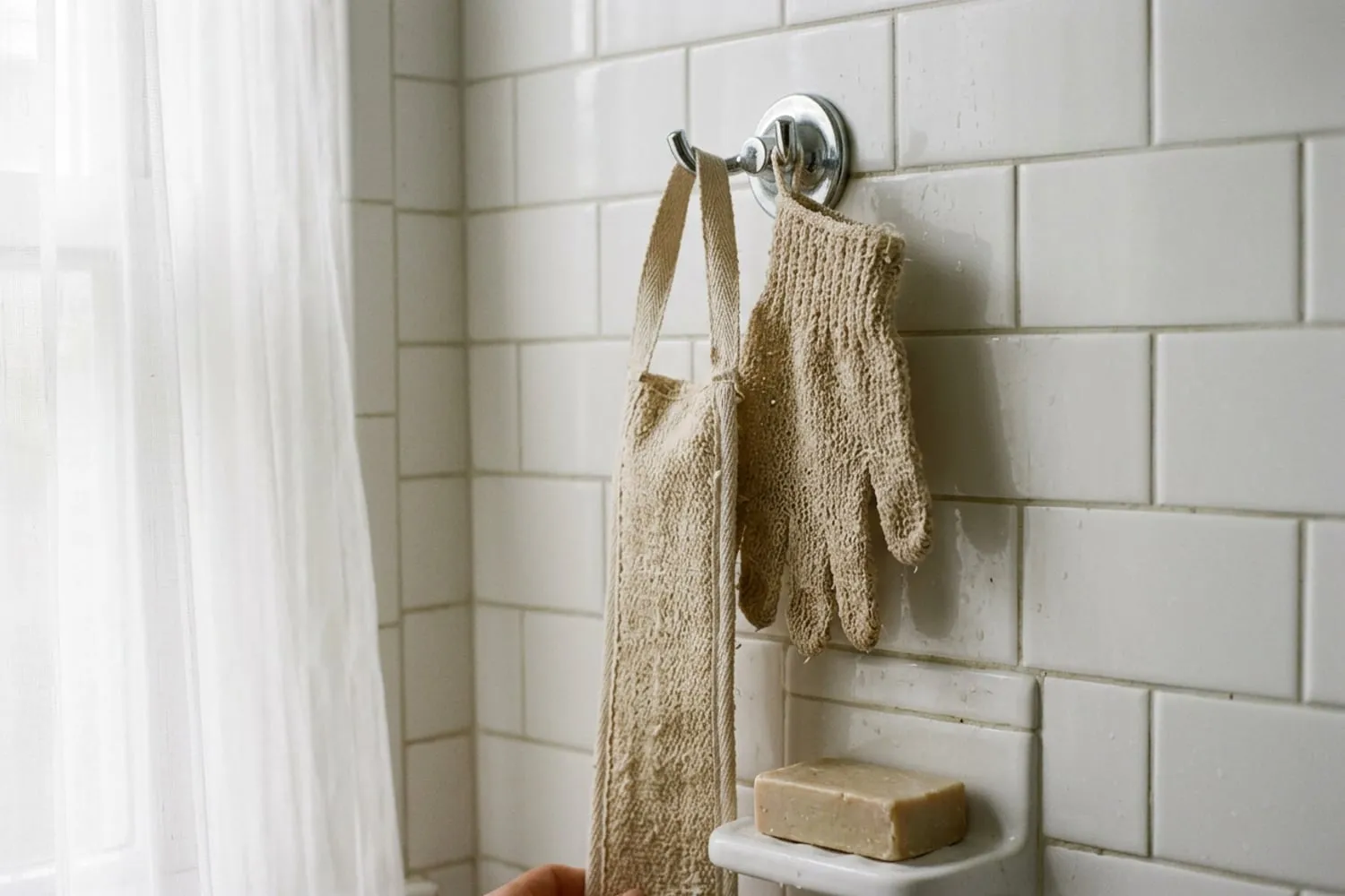 A damp textured exfoliating glove and a long cloth back-scrub strap hanging side by side on a chrome hook in a white-tiled shower, a bar of plain soap on a ledge below