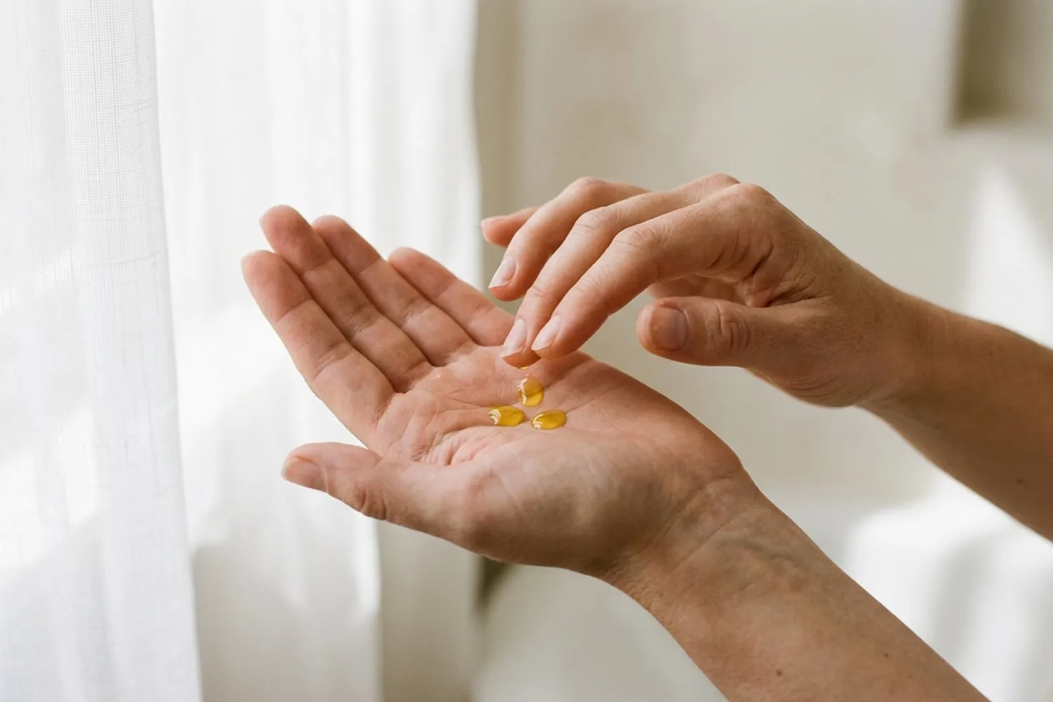 A close view of three small drops of golden oil in a cupped palm, the other hand’s fingertips about to touch them, against a soft-focus background of a white bathroom