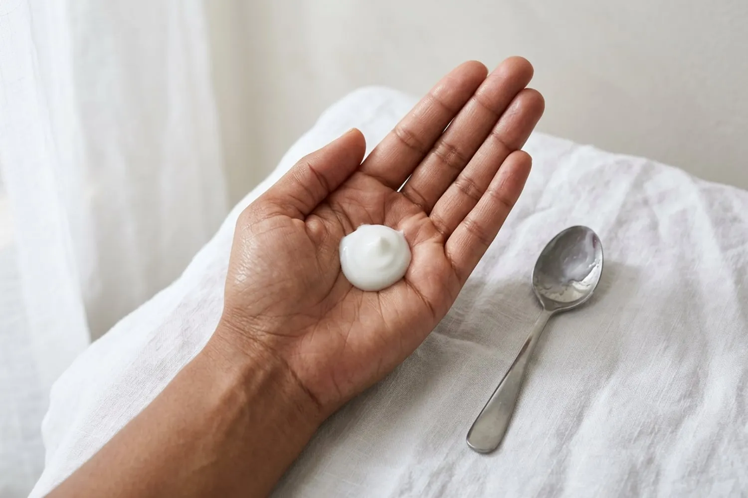 A full teaspoon of white sunscreen lotion tipped into an open palm beside a small steel teaspoon, seen from above on a white linen towel; medium brown skin in soft morning light