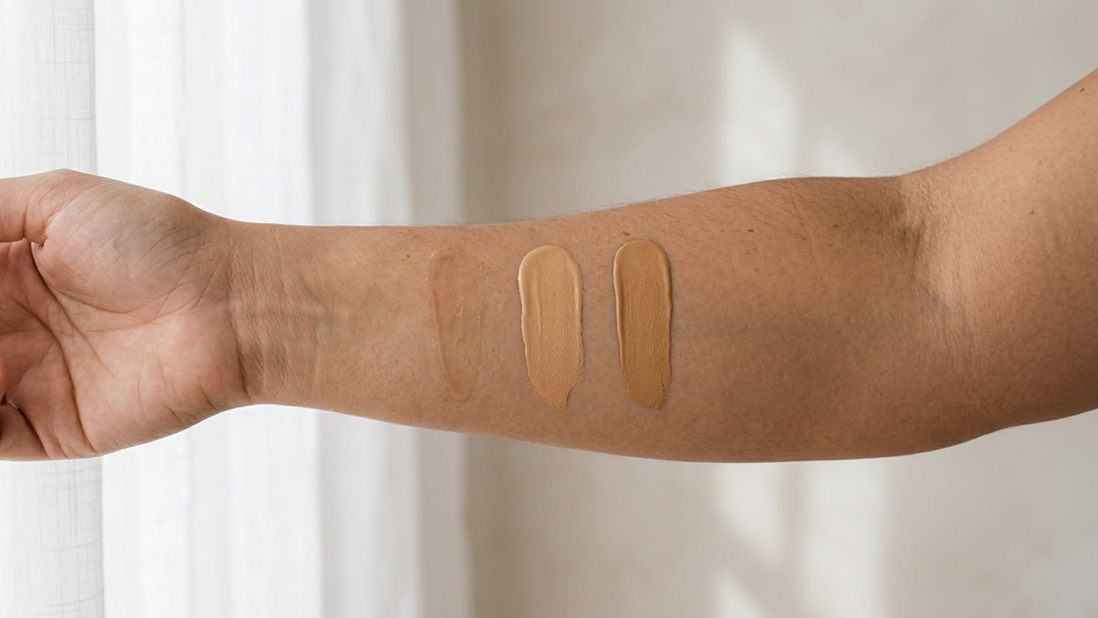 Three swatches in a row on an inner forearm, from a sheer, watery skin tint to a soft tinted moisturizer to an opaque stripe of foundation; warm medium-tan skin in window light.