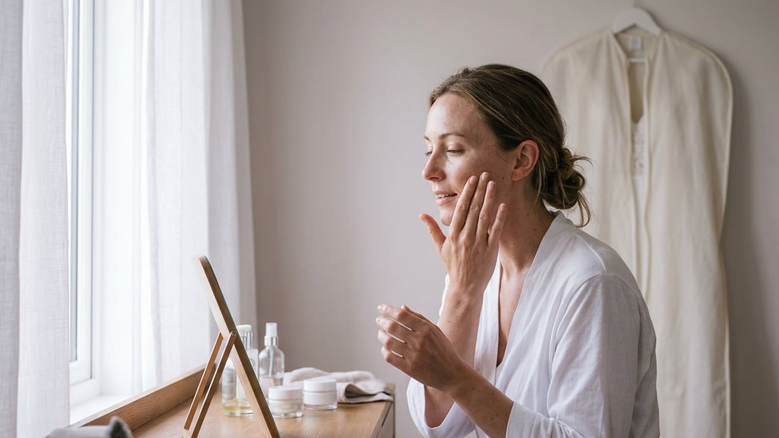 A woman in her late twenties smoothing moisturizer onto her cheek at a dressing table by a window, a wedding dress in a garment bag hanging on the wall behind her; soft morning light.