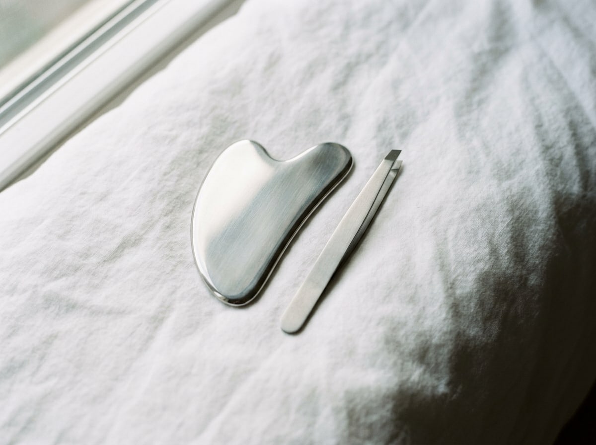 A stainless steel gua sha and a pair of slant-tip tweezers on white cotton.